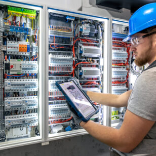 A man in a hard hat uses a tablet while manually inspecting a switchboard. A balance of digital diagnostics and practical action, with artificial lighting and technical precision.